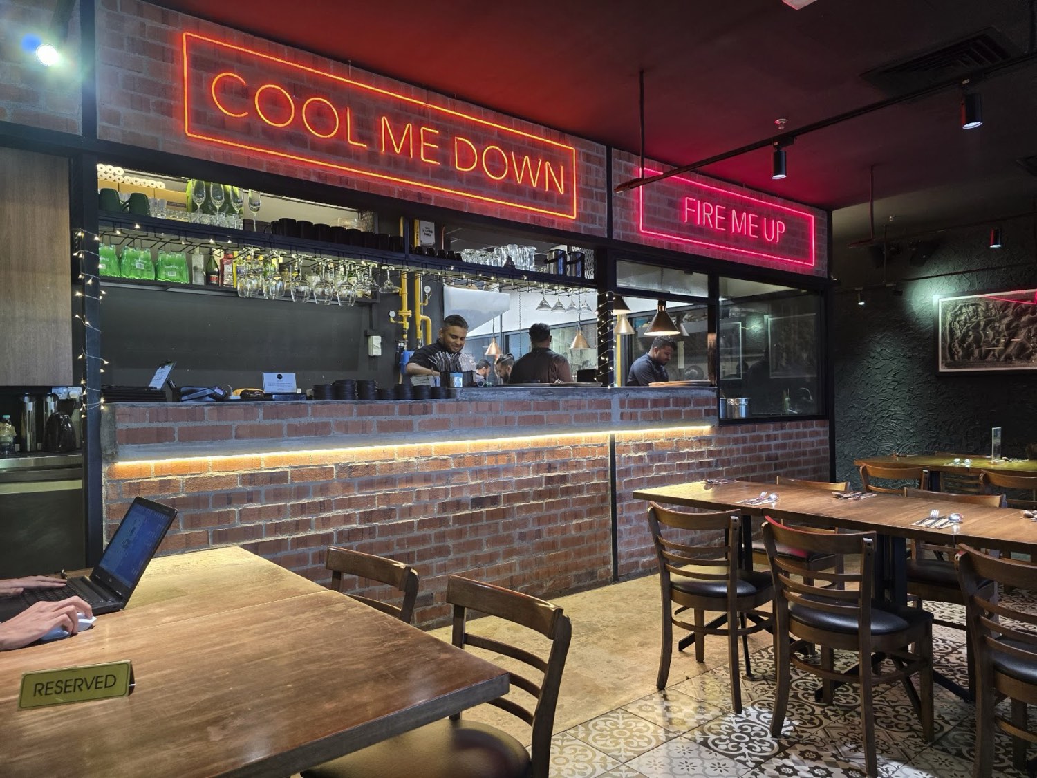 The open kitchen pass at Fire By Shankar under neon &lsquo;Cool Me Down&rsquo; and &lsquo;Fire Me Up&rsquo; signs, against exposed brick